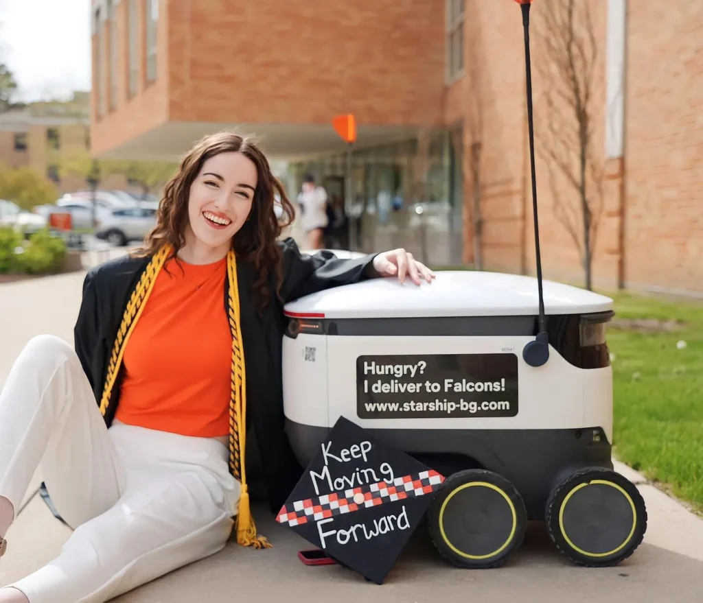 Graduate smiling at the camera next to a Starship robot