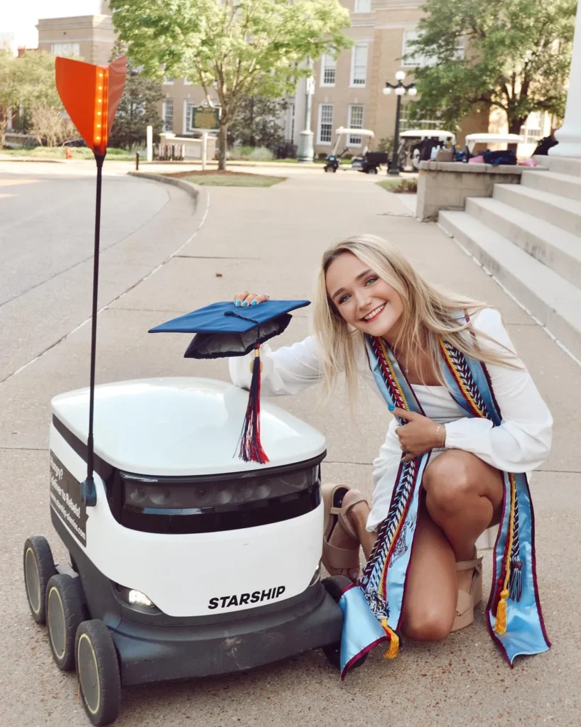 Graduate smiling at the camera next to a Starship robot