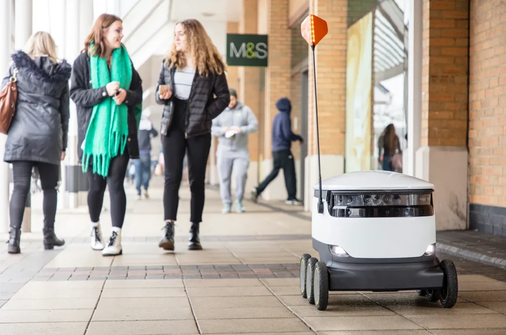 A Starship robot rolling on the street with pedestrians